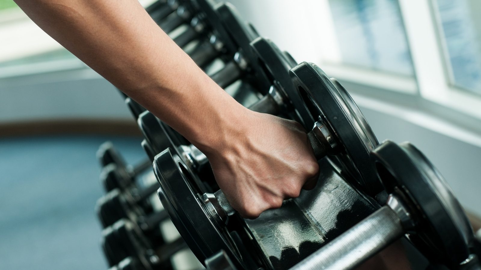 A hand reaching for a dumbbell on a rack in a gym lit by a window