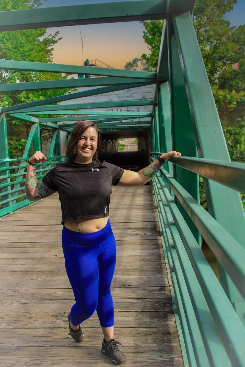 Jess standing on a green footbridge at golden hour, flexing one arm and laughing
