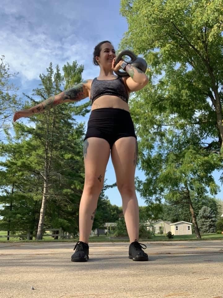 Jess standing outdoors with a kettlebell racked on one shoulder, smiling