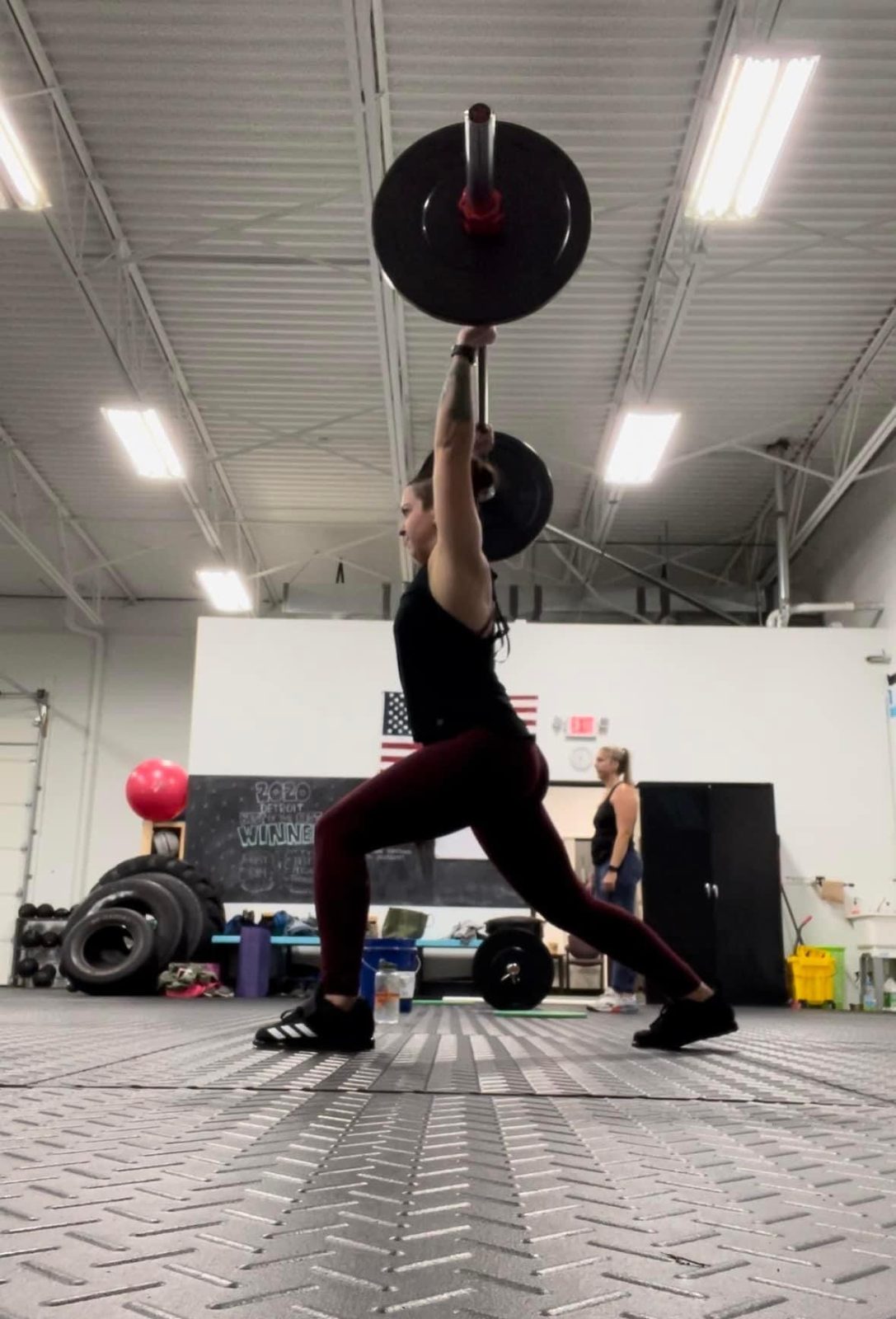 Jess pressing a loaded barbell overhead in a split stance at the gym