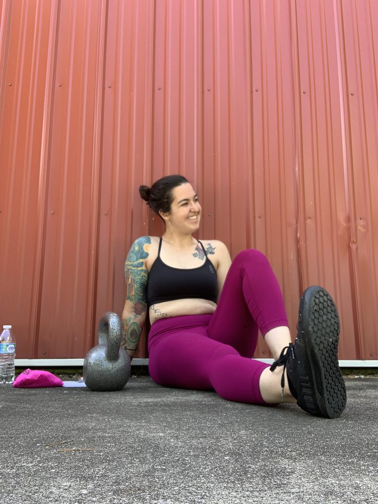 Jess sitting on the ground against a red metal wall after training, laughing, a kettlebell beside her