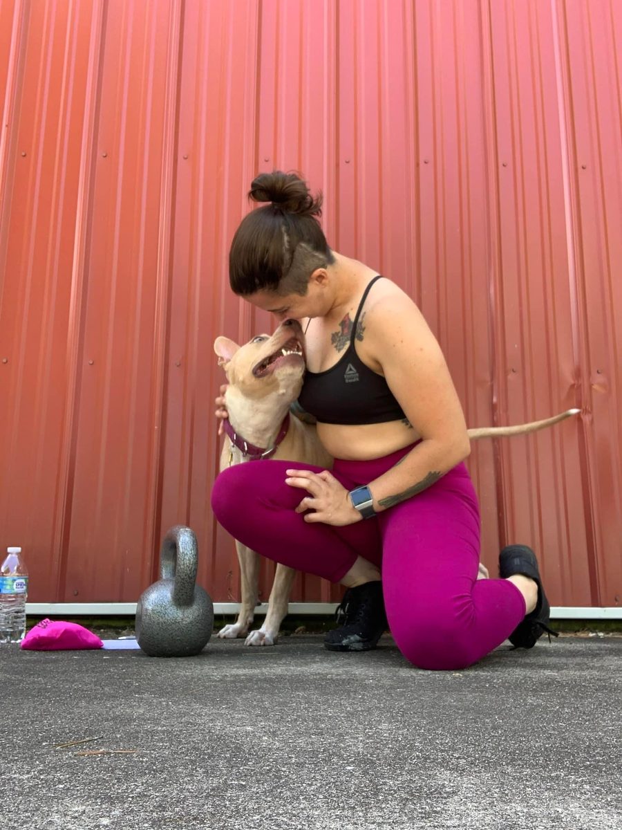 Jess kneeling to greet her dog nose to nose against a red metal wall, a kettlebell on the ground beside them