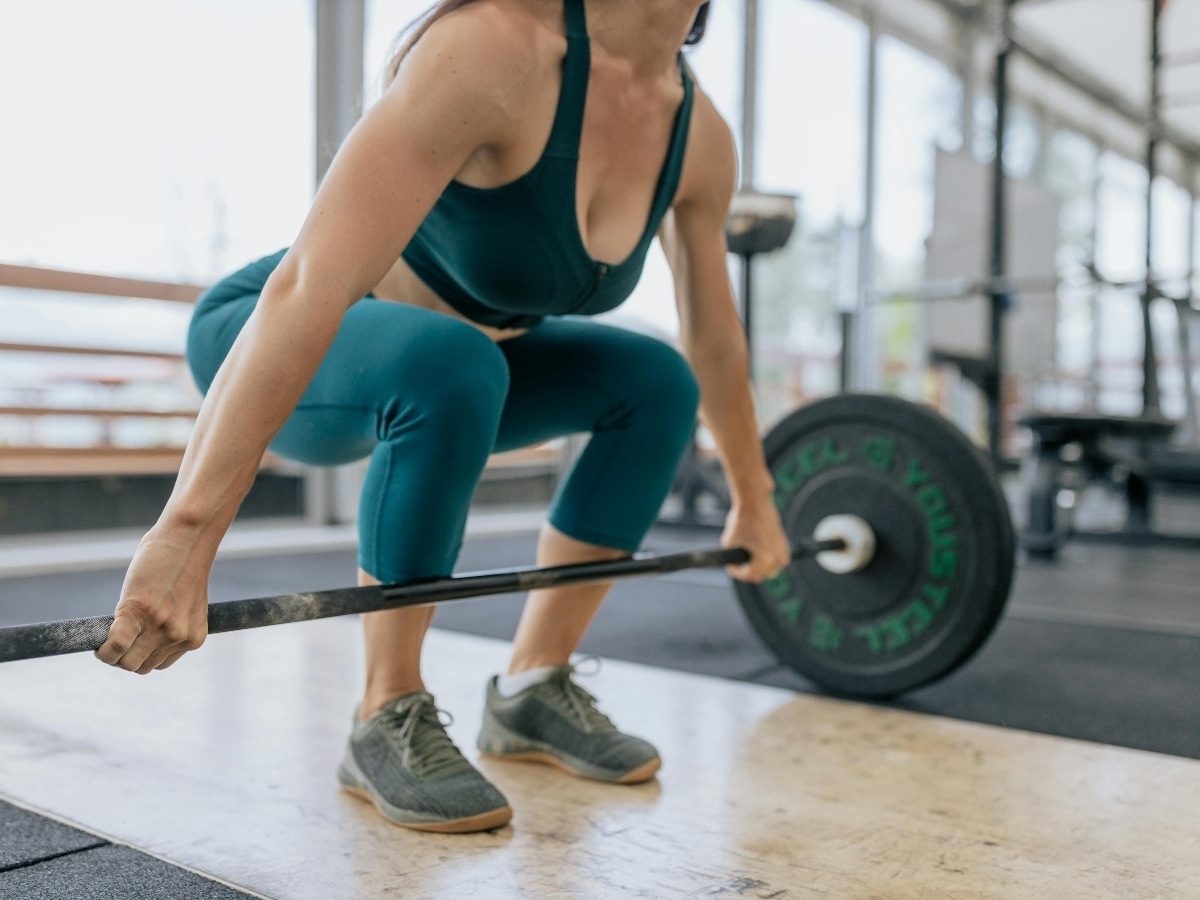 Woman in teal setting up a barbell deadlift in a bright gym